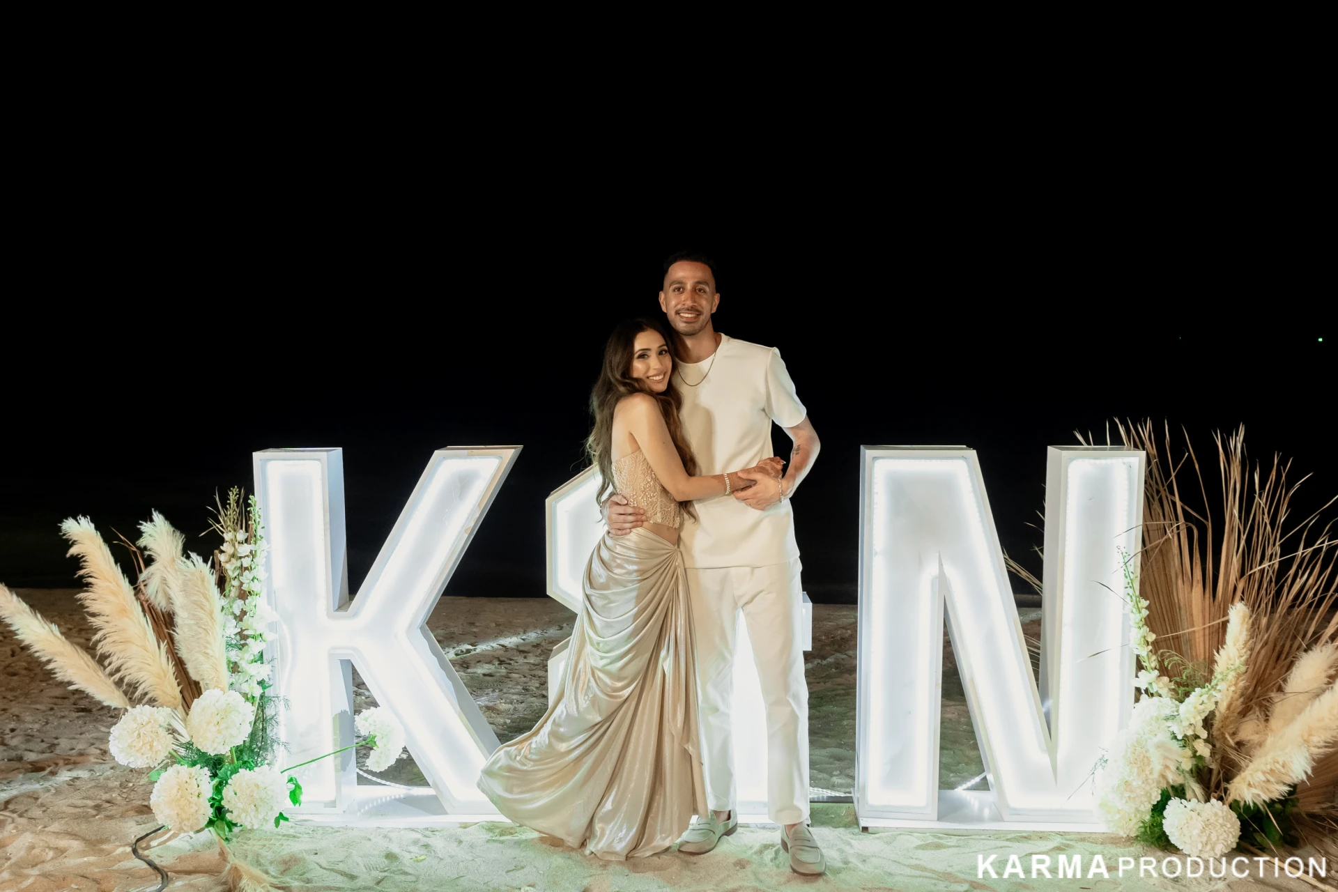 Kavya and Nikhil beside their illuminated monogram on the beach